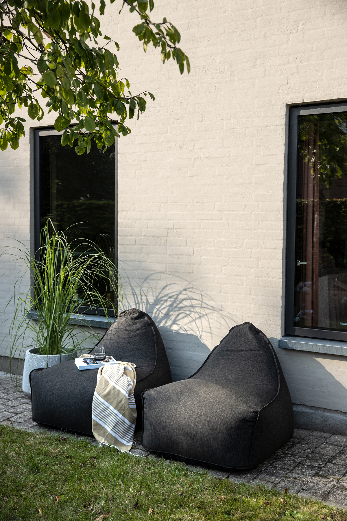To mørkegrå sækkestole på en terrasse ved en lys murstensvæg, med stribet plaid, bog og glas ved siden af en potteplante i sollys.