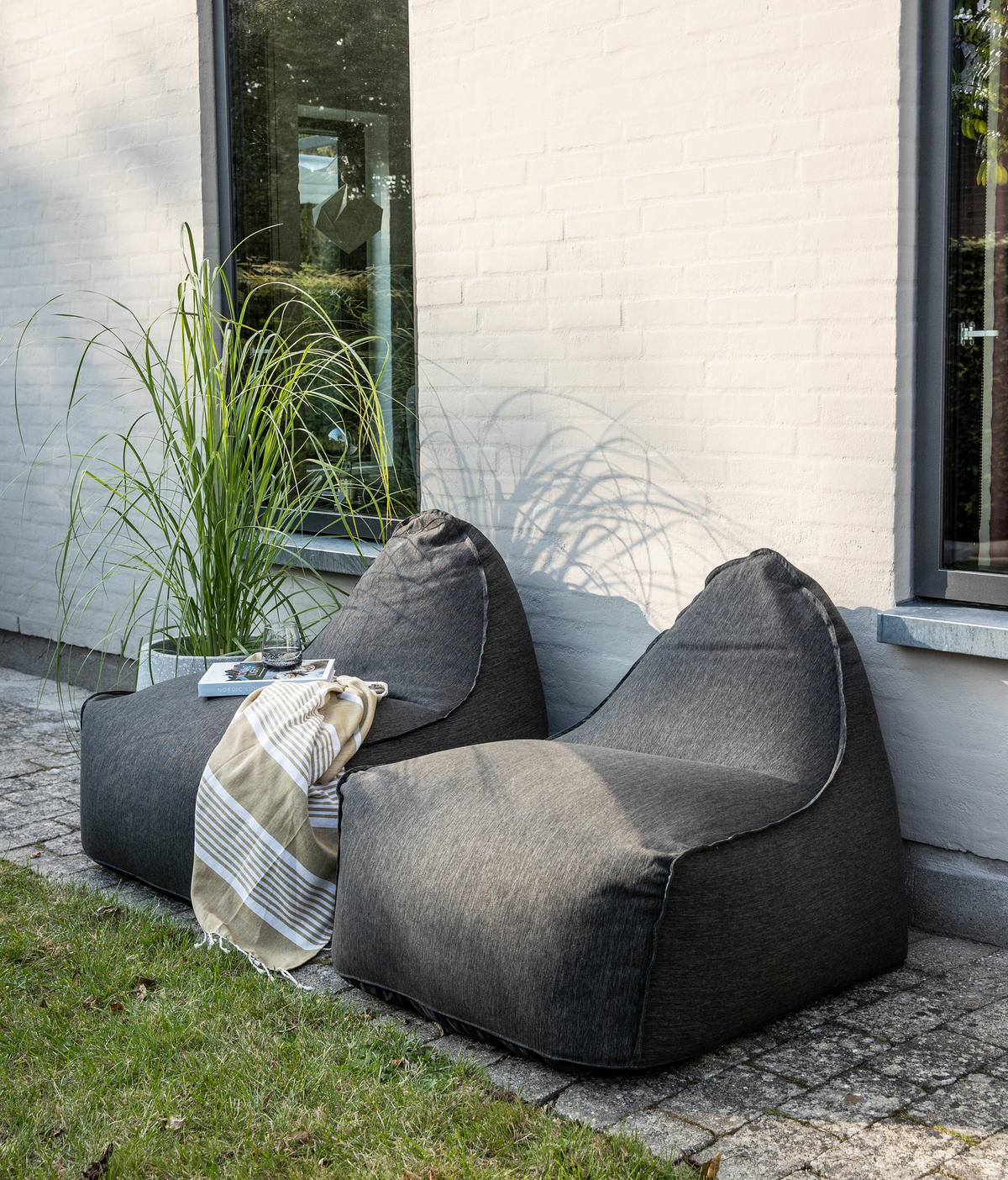 To mørkegrå sækkestole på en brolagt terrasse ved en hvid mur, med prydgræs, bog, glas og stribet plaid i dagslys.