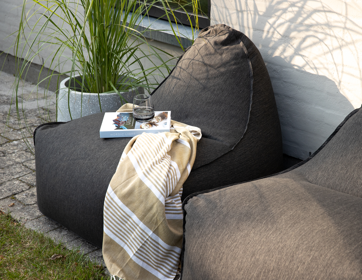 To mørkegrå sækkestole udendørs med et beige stribet tæppe, et glas vand og bogen ”Nordic Light” på en brolagt terrasse ved siden af en potteplante.