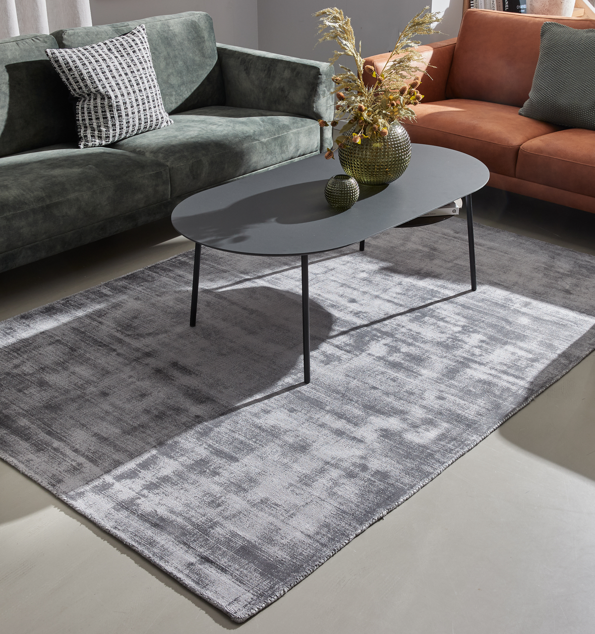 Gråt rektangulært tæppe i stuen med mørkegråt ovalt sofabord mellem en grøn sofa og en cognacfarvet sofa, pyntet med vase og tørrede kviste.