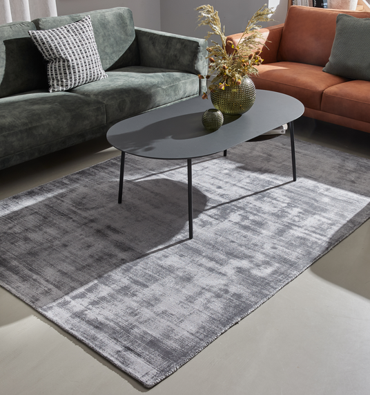 Gråt rektangulært tæppe i stuen med mørkegråt ovalt sofabord mellem en grøn sofa og en cognacfarvet sofa, pyntet med vase og tørrede kviste.