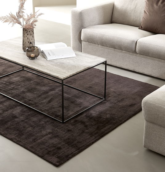 Beige sofa og lænestol med mørkebrun tæppe og rektangulært sofabord med stenplade og sort metalstel; vase med tørrede grene, lille lyslygte og en opslået bog på bordet.