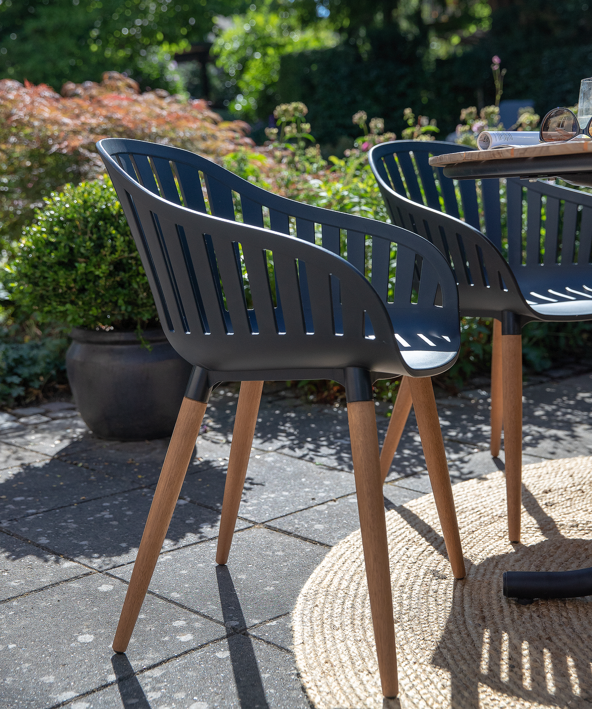 Sort udendørs stol med lamel-ryg og træben ved rundt bord på flisebelagt terrasse, solskin og grøn beplantning i baggrunden.