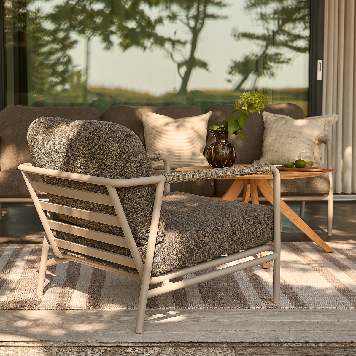 Udendørs lounge med beige metallarmlænestol, sofa og lille træbord med brun glasvase med hortensia, puder og stribet tæppe på en terrasse ved skydedøre.