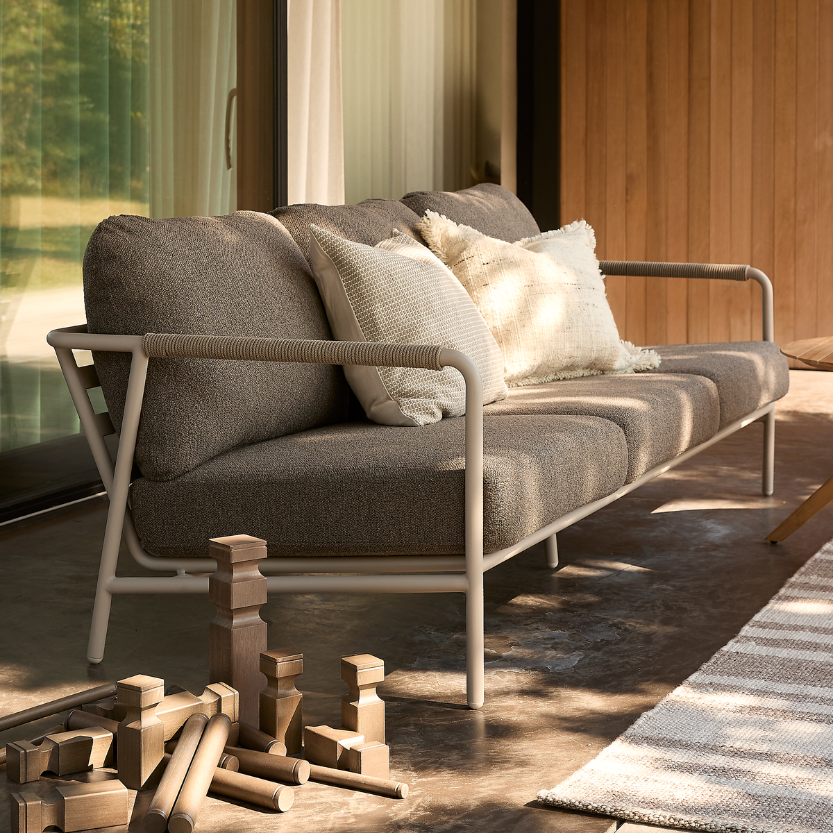 Udendørs sofa med grå puder og lys metalramme, neutrale pyntepuder; solbeskinnet terrasse ved skydedøre, træklodser på betongulv og et stribet tæppe.