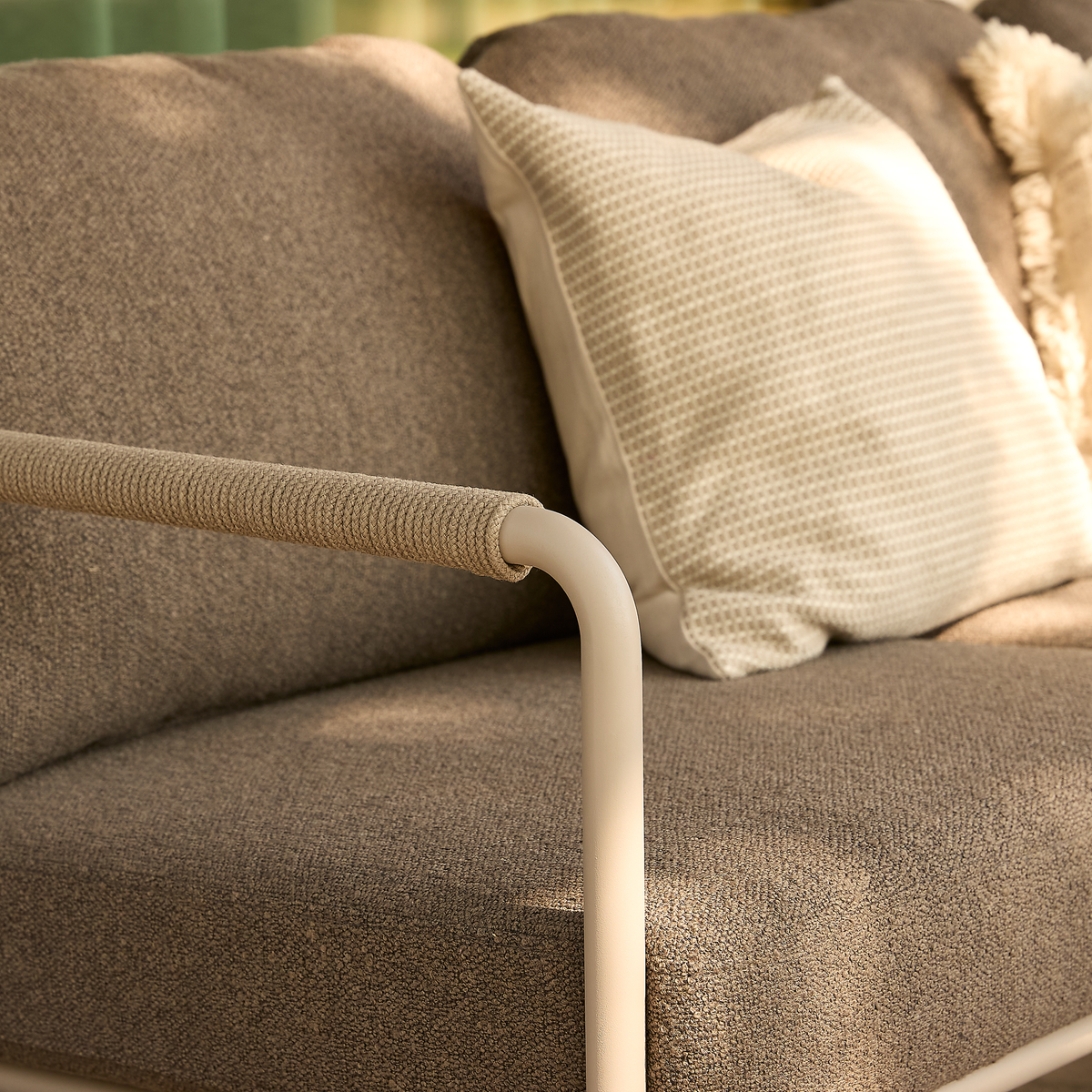 Nærbillede af beige sofa med tekstureret pude og omviklet metalarm i blødt dagslys.