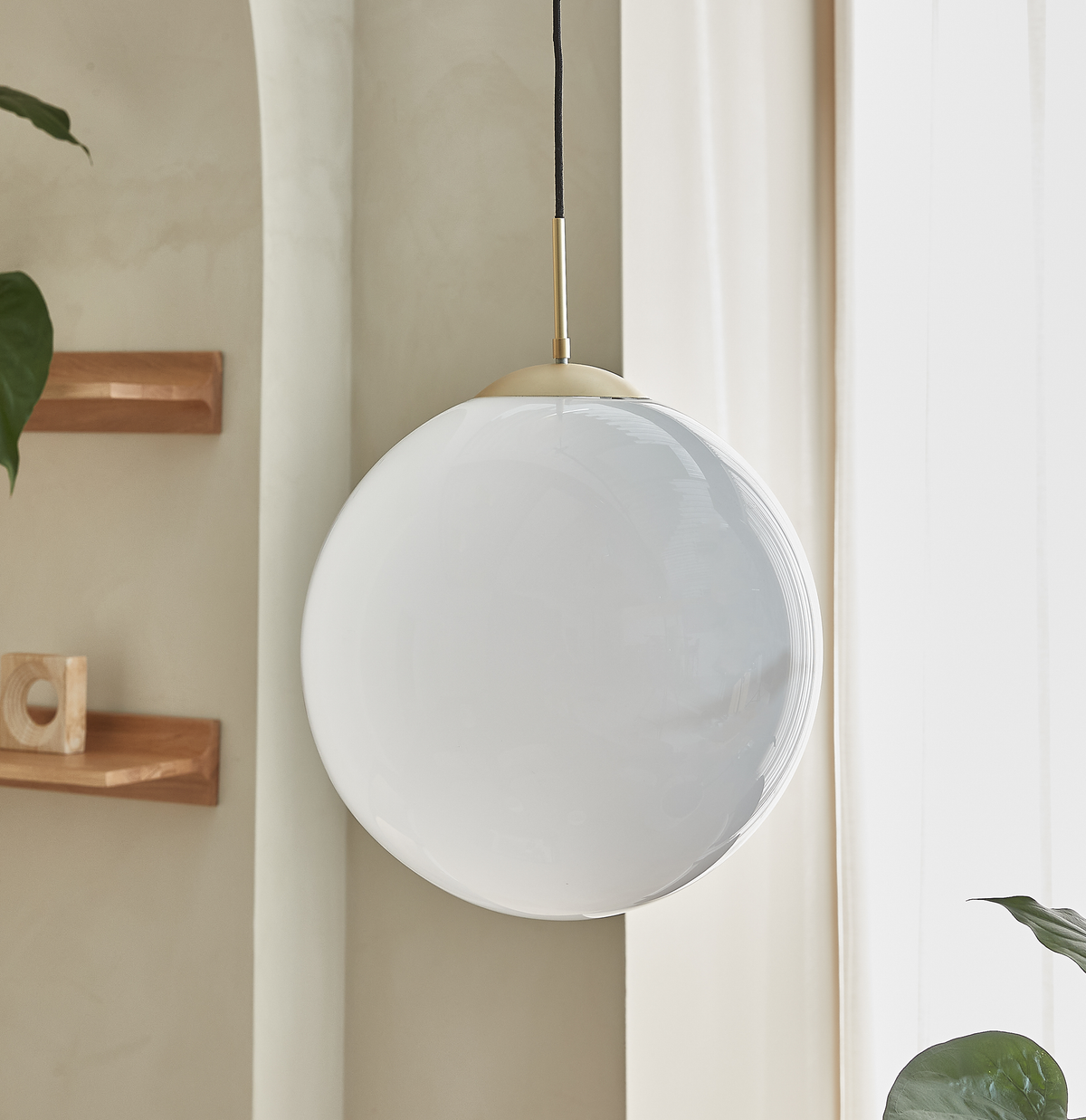 Hvid glaskugle‑pendel hænger ved vindue i lys, minimalistisk indretning med træhylder og grøn plante.