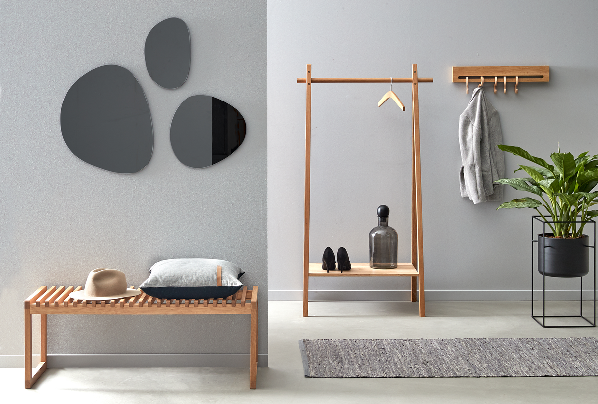 Minimalistisk entré med grå vægge: træbænk med pude og hat, tøjstativ og knager med jakke, organiske spejle, løber og grøn plante.