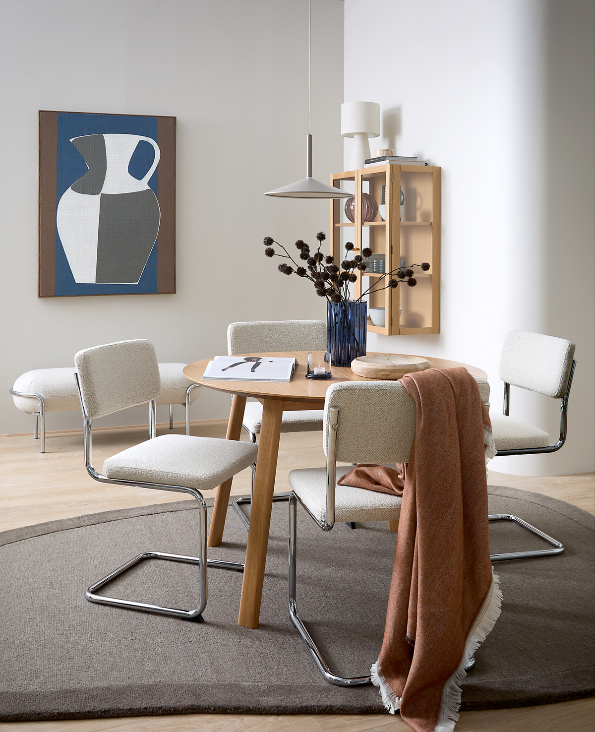 Lyst skandinavisk spisekrog med rundt egetræsbord, fire cremefarvede cantilever-stole med krom, en rustfarvet plaid over en stol, blå vase med tørrede grene, pendel og vægskab; på bordet ligger bøger og en side med bogstavet A; grafisk plakat med kande på væggen.