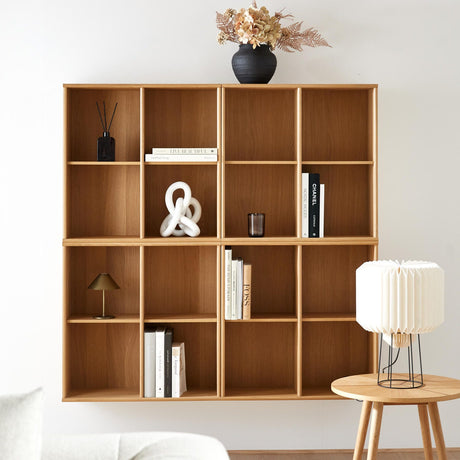 Væghængt reol i træ med bøger, CHANEL‑bog, skulpturel hvid knude, sort vase med tørrede blomster samt en lille bordlampe og en plisseret bordlampe på et sidebord.