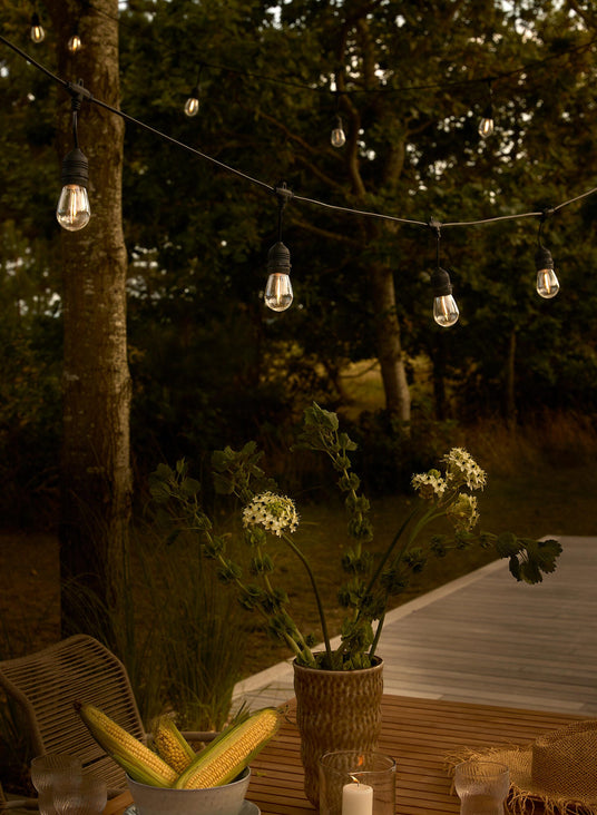 Hygge på terrassen i skumringen med en varm lyskæde over et træbord, vase med hvide blomster, majskolber i en skål og tændt bloklys.