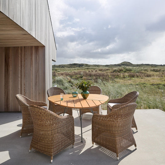 Terrasse med rundt træbord og seks kurvestole; vase med grønne grene og to glas på bordet; udsigt til græsklædte klitter i dagslys.