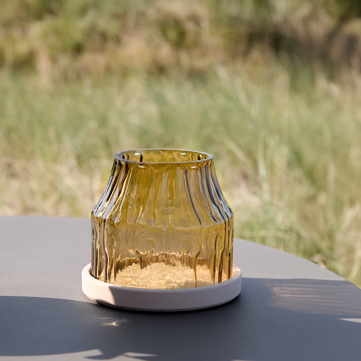 Ravfarvet rillet glaslyslygte/vase på rund lys bakke på udendørs bord i dagslys.