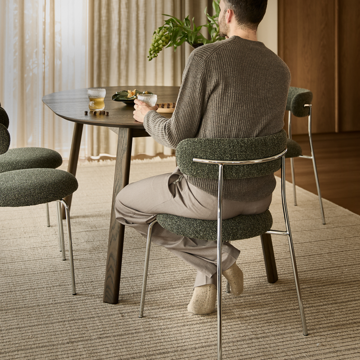 Mand sidder ved rundt spisebord med grønpolstrede stole, glas med drik og snacks i blødt dagslys i spisestue.