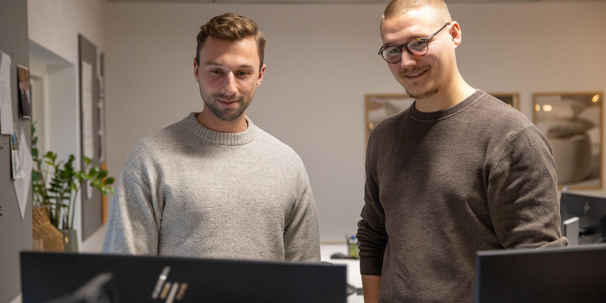 To personer står bag computerskærme på et kontor; HP‑logo på skærmen i forgrunden.