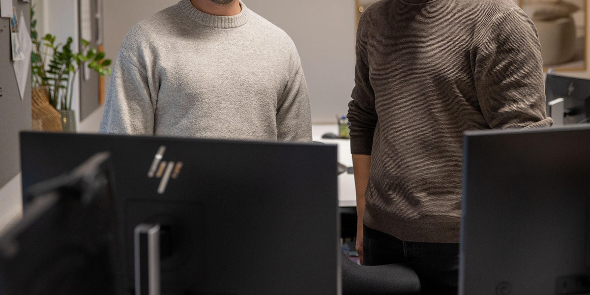 To personer står bag computerskærme på et kontor; HP‑logo på skærmen i forgrunden.