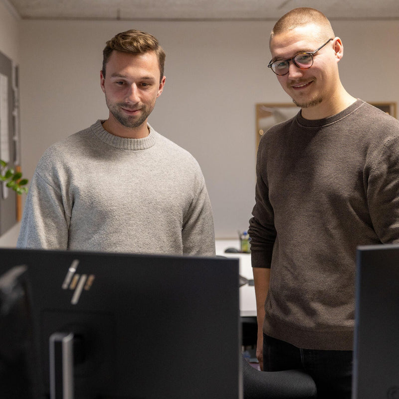 To personer står bag computerskærme på et kontor; HP‑logo på skærmen i forgrunden.
