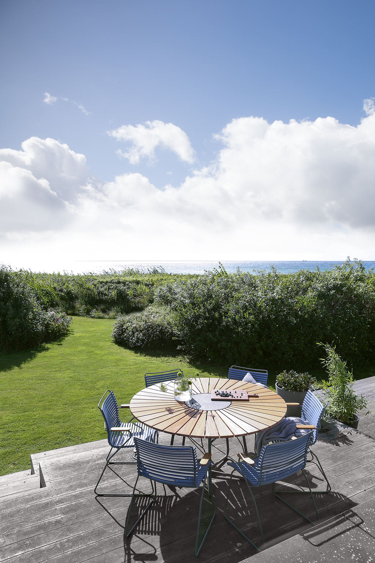 Rundt udendørs træbord med blå stole på terrasse, udsigt over græsplæne og hav under blå himmel med skyer.