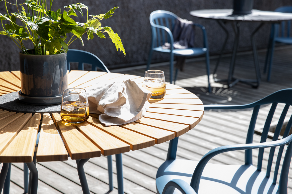 Rundt træbord med blå stole på terrasse i sollys; to glas med is og drik, en potteplante og en stofserviet på bordet.