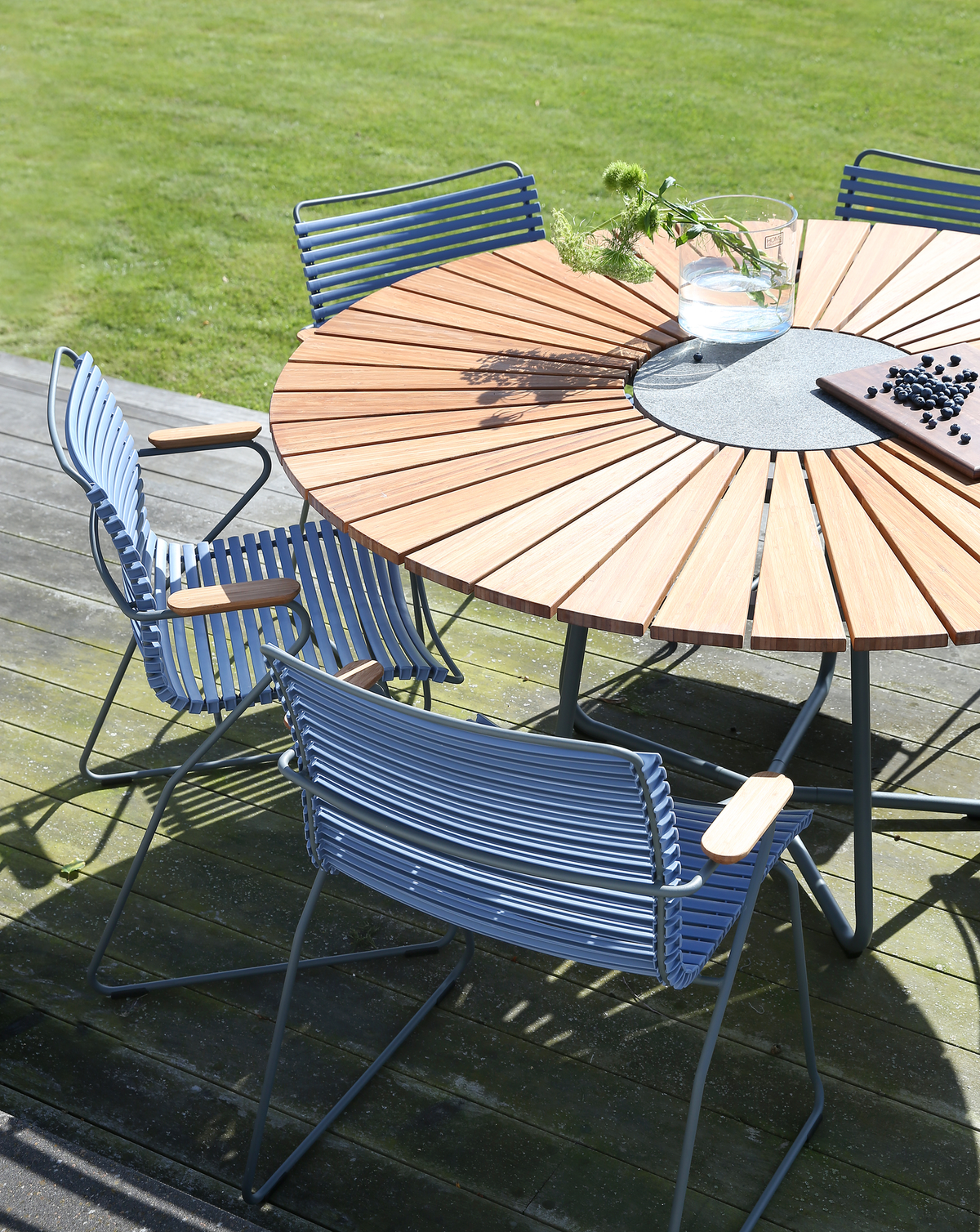 Rundt havebord i træ med stencenter, omgivet af blå metalstole på en solrig terrasse; glasvase med vand og grønne kviste samt skærebræt med blåbær.