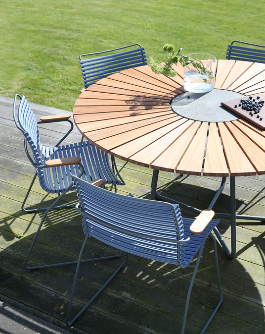 Rundt havebord i træ med stencenter, omgivet af blå metalstole på en solrig terrasse; glasvase med vand og grønne kviste samt skærebræt med blåbær.