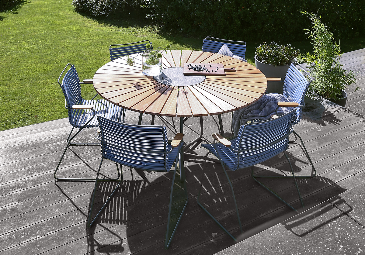 Rundt havebord i træ med blå stole på terrasse; vase med grøn kvist, skærebræt med bær og en plaid over en stol i solrig have.