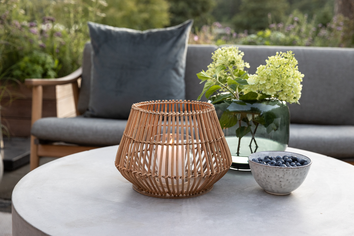 Udendørs sofabord med rottinglanterne med tændt lys, grøn glasvase med hortensia og en skål med blåbær foran en grå sofa.