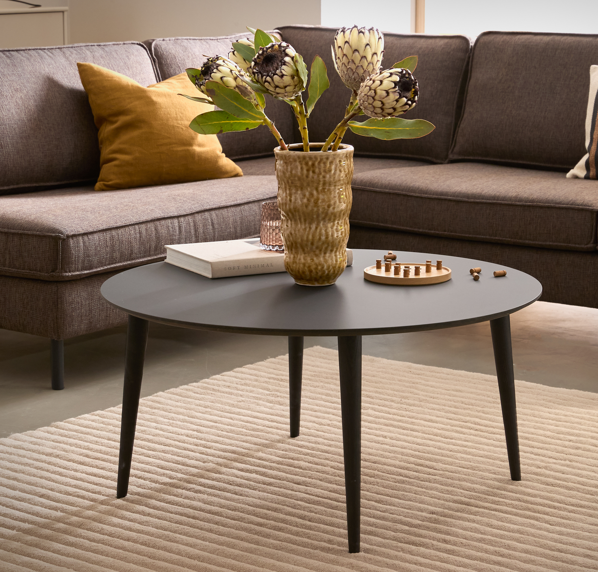 Rundt sort sofabord med vase med protea, bogen Soft Minimal og et lille træspil foran en brun sofa på et beige rillet tæppe.