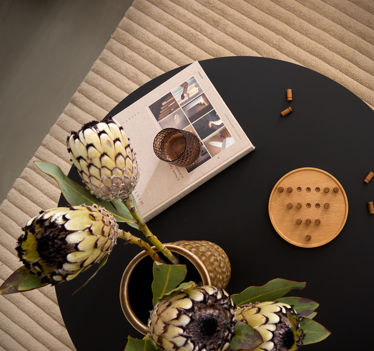 Rundt sort sofabord på beige rillet tæppe med protea i messingvase, en bog med interiørbilleder, et lille glas og et rundt træspil med pinde.