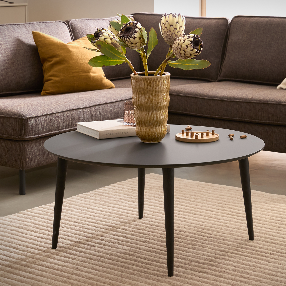 Rundt sort sofabord på beige tæppe med vase med protea, en bog og et lille brætspil i en moderne stue med brun sofa og sennepsgul pude.