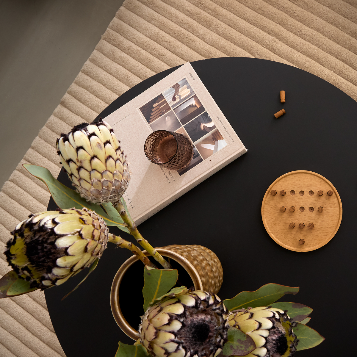 Set oppefra: rundt sort sofabord med protea i messingvase, beige bog, brunt glas og et lille træspil på et rillet beige tæppe.