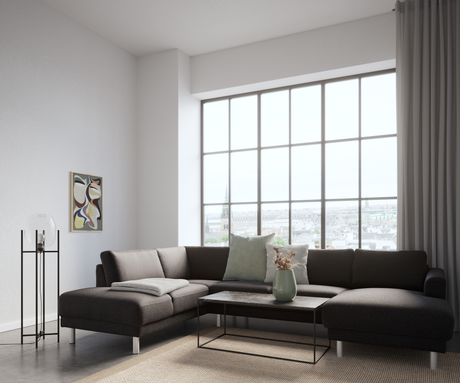 Lys stue med mørk hjørnesofa foran store sprossede vinduer med byudsigt, sofabord i sten med grøn vase og tørrede grene, grå puder og plaid, gulvlampe og abstrakt vægkunst.