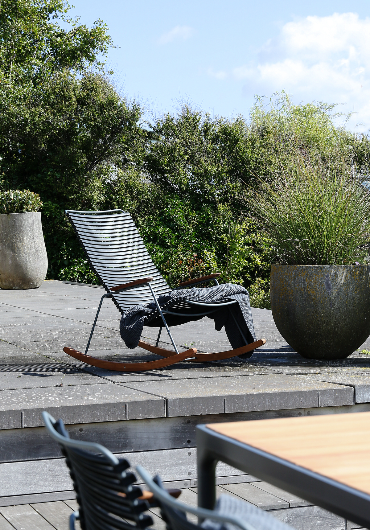 Moderne gyngestol i metal og træ på terrasse med plaid og store krukker med prydningsgræs i solskin.