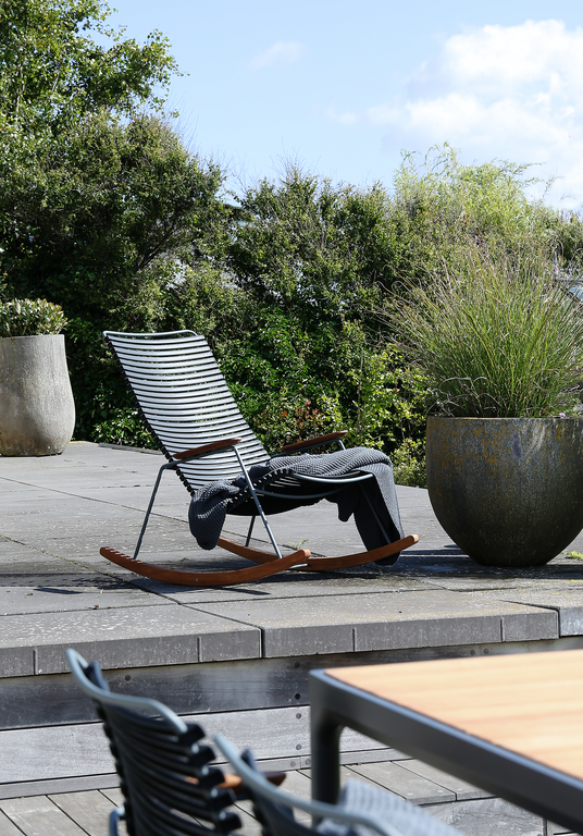 Moderne gyngestol i metal og træ på terrasse med plaid og store krukker med prydningsgræs i solskin.