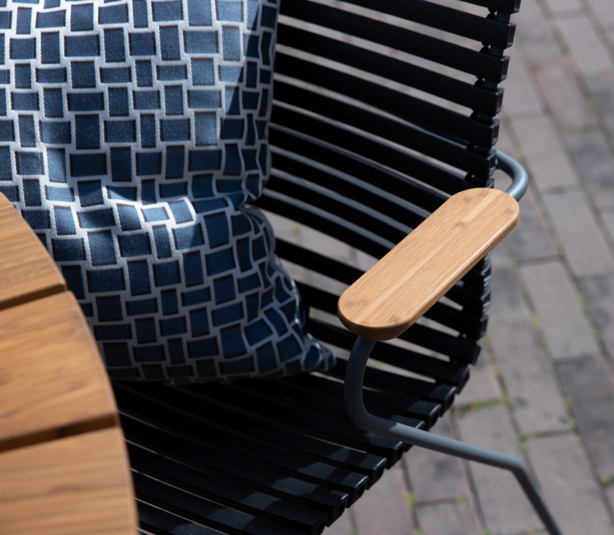 Nærbillede af en moderne sort udendørsstol med armlæn i træ og en blåmønstret pude ved siden af en rund bordplade i træ på en brolagt terrasse.