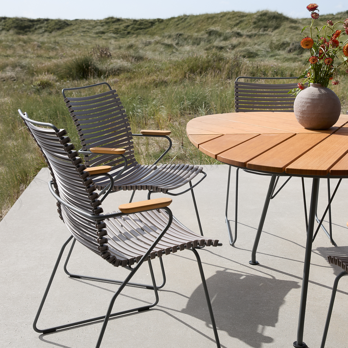 Rundt udendørs bord i træ med metalben og lamelstole i metal med træarmlæn på en terrasse ved klitter; vase med blomster på bordet.
