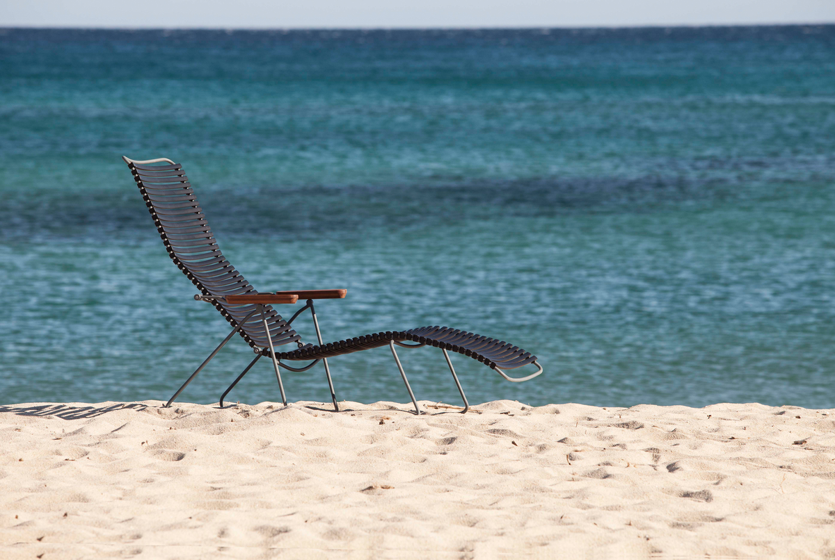 En moderne liggestol i metal og træ står på en sandstrand med turkisblåt hav i baggrunden.