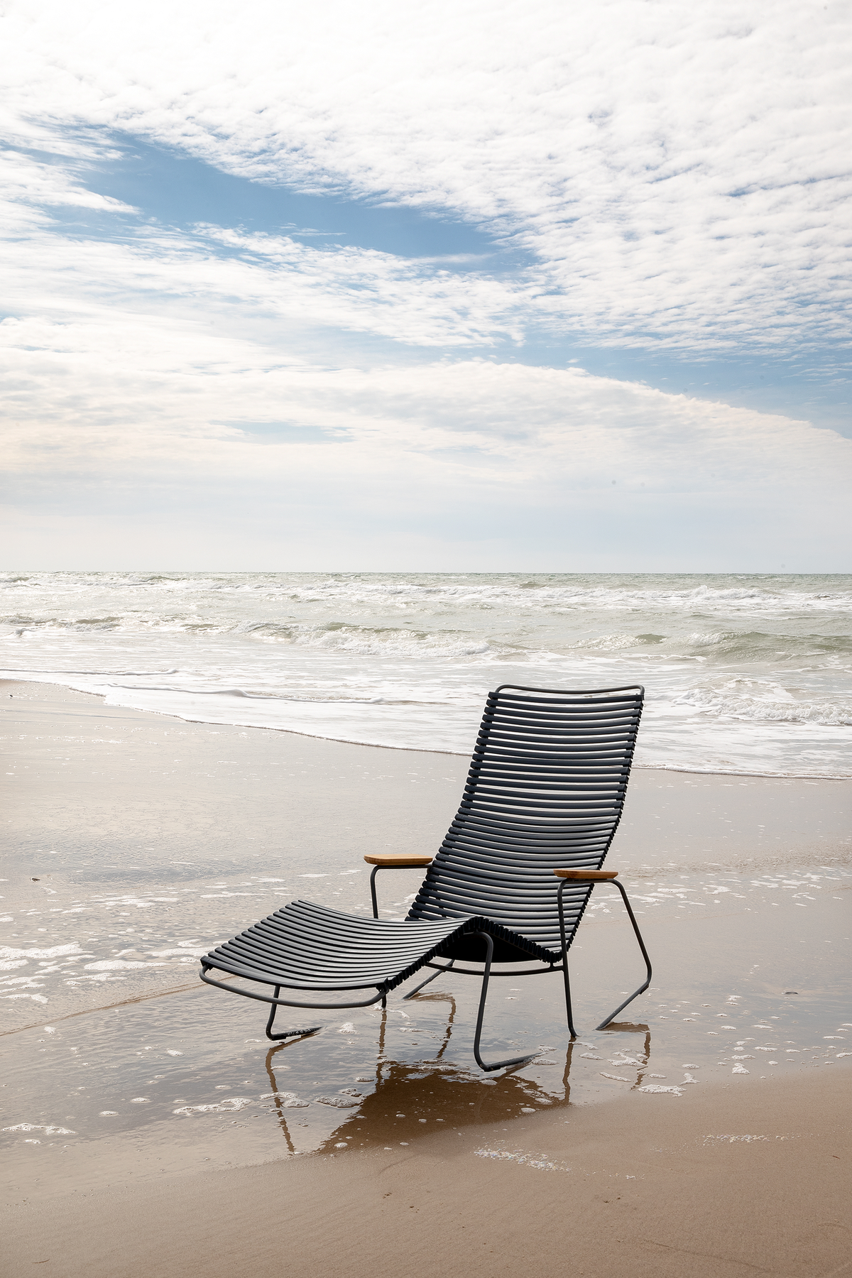 En sort lamel-liggestol med armlæn står på en sandstrand ved havet, med bølger og lys skyet himmel.