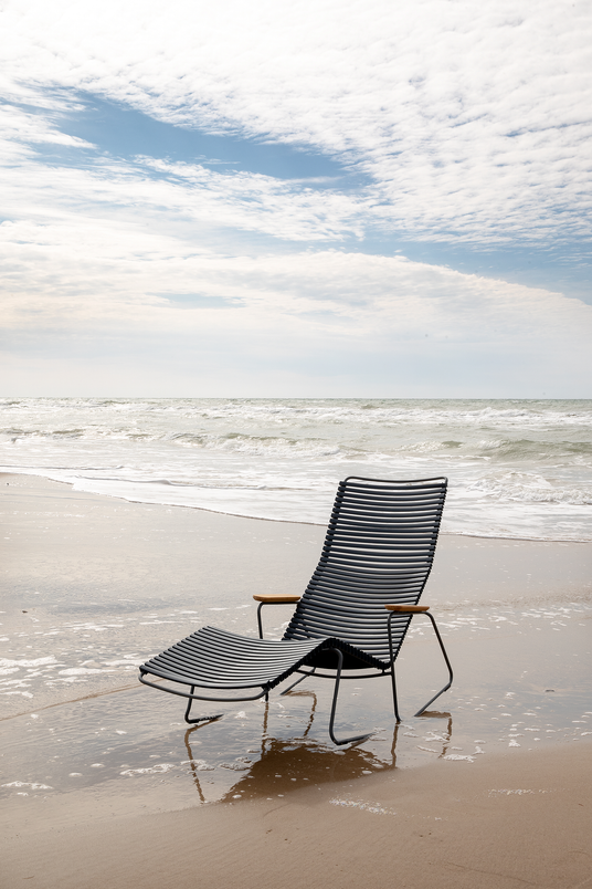 En sort lamel-liggestol med armlæn står på en sandstrand ved havet, med bølger og lys skyet himmel.