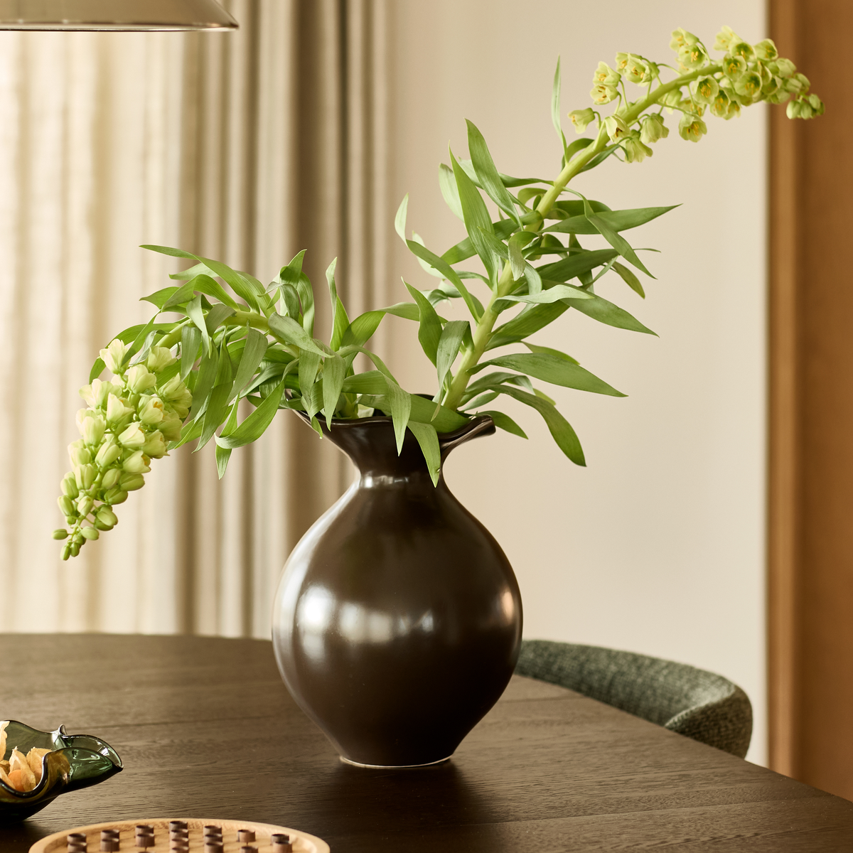 Mørk keramisk vase med grønne grene på et mørkt spisebord i en lys, skandinavisk indretning.