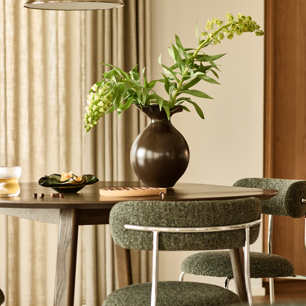 Mørkebrun vase med grønne grene på rundt spisebord i træ, grønpolstrede stole samt glas og skål med citrus i en varm, lys spisestue med gardiner.
