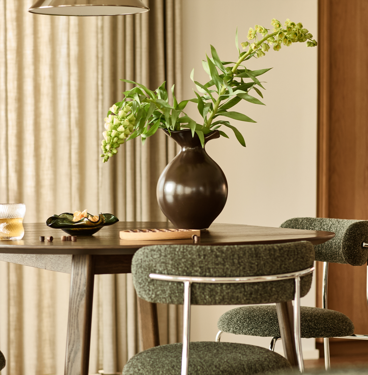 Rundt spisebord i mørkt træ med grønne polstrede stole, brun vase med grønne grene, skål og glas i et lyst, roligt spiseområde.