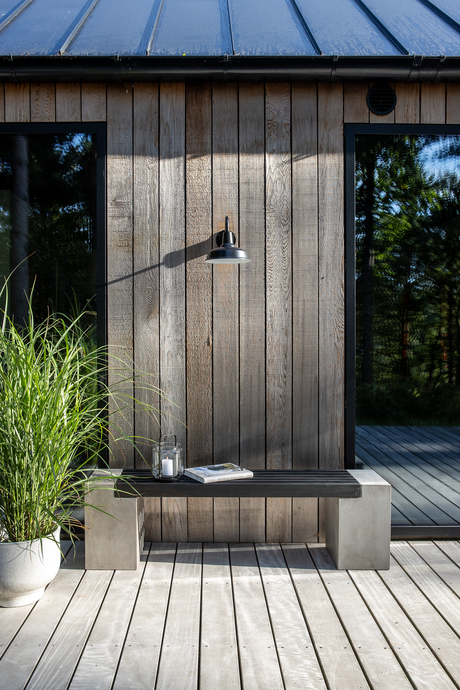 Facade i vejrgråt træ med sort væglampe over minimalistisk bænk på terrasse; potteplante, lanterne med lys og en bog på sædet.