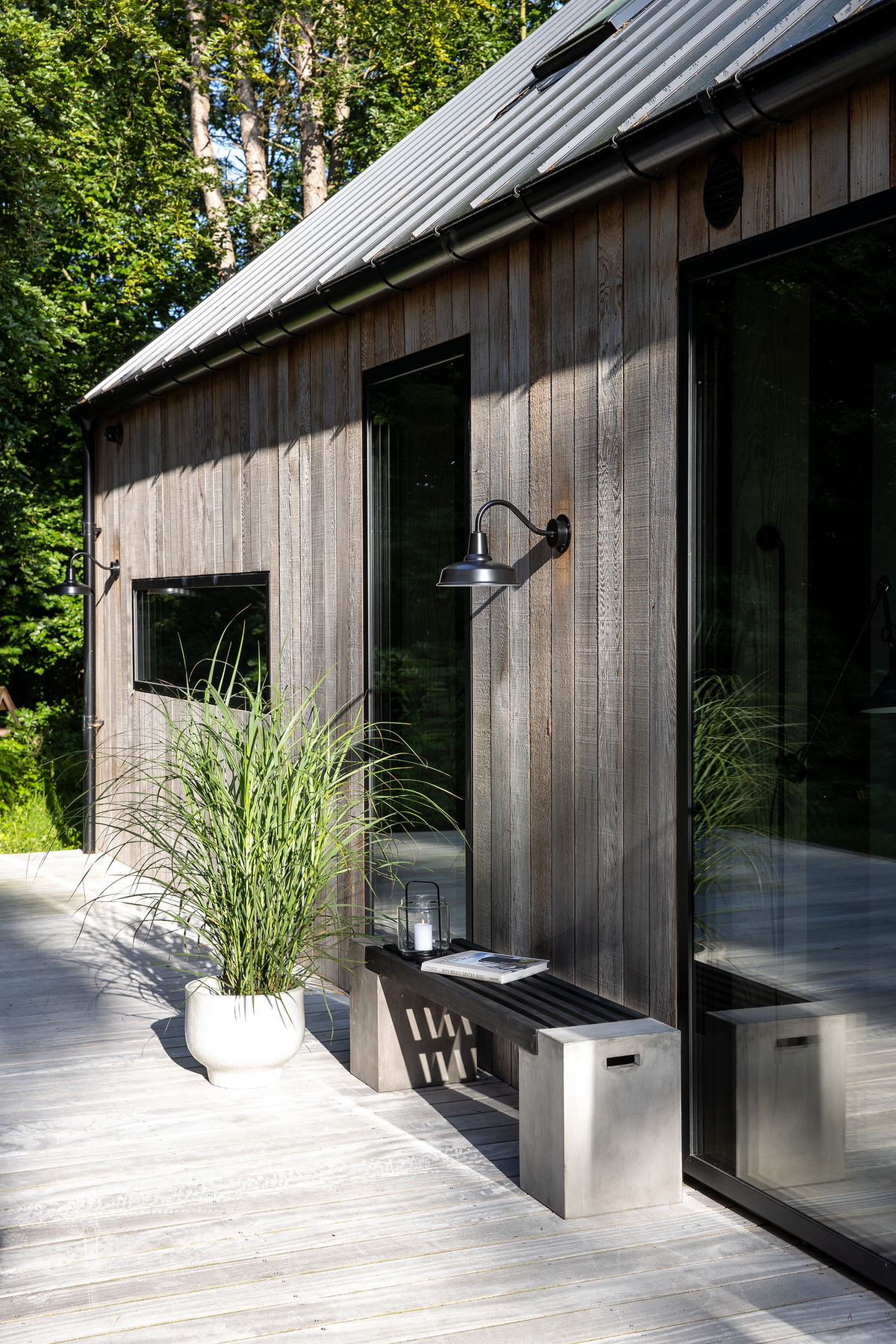 Træfacade med store glaspartier, sort væglampe og en bænk med lanterne og blad på en lys træterrasse, krukkeplante ved siden af; solrigt ved skov.