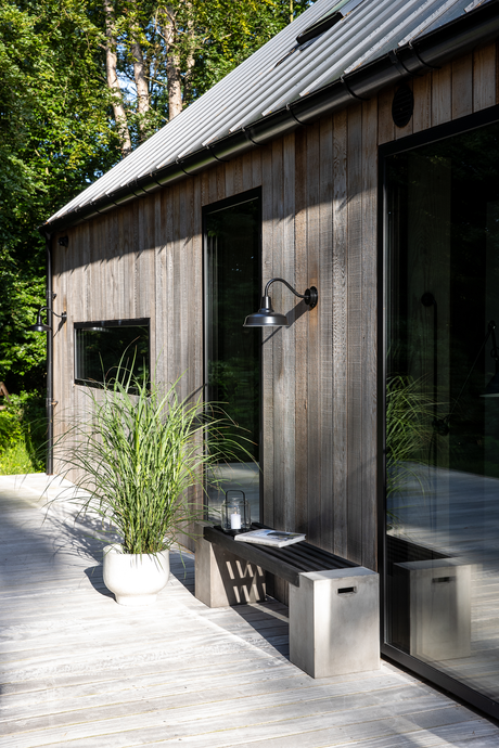 Træfacade med store glaspartier, sort væglampe og en bænk med lanterne og blad på en lys træterrasse, krukkeplante ved siden af; solrigt ved skov.