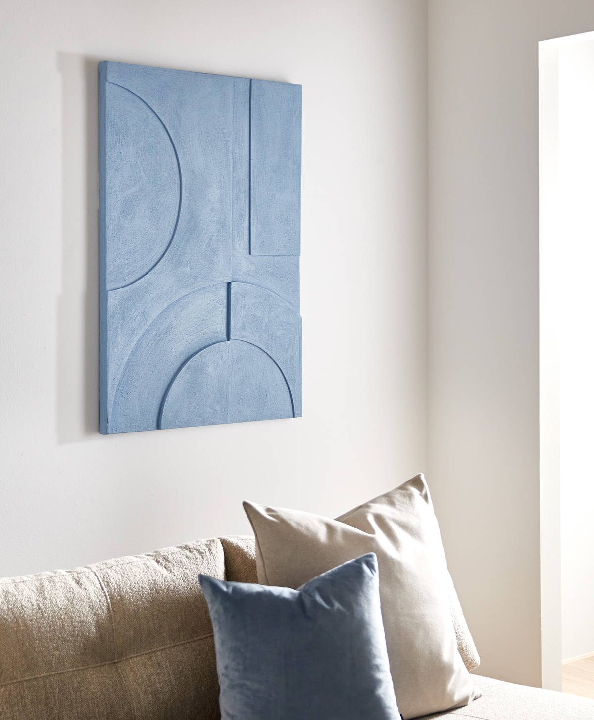 Blå geometrisk relief-vægkunst over en beige sofa med blå og beige puder i et lyst stueområde.