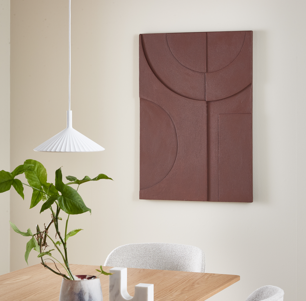 Spiseområde med hvid plisseret pendel, grøn plante i vase på træbord og brun geometrisk vægpanel på lys væg.