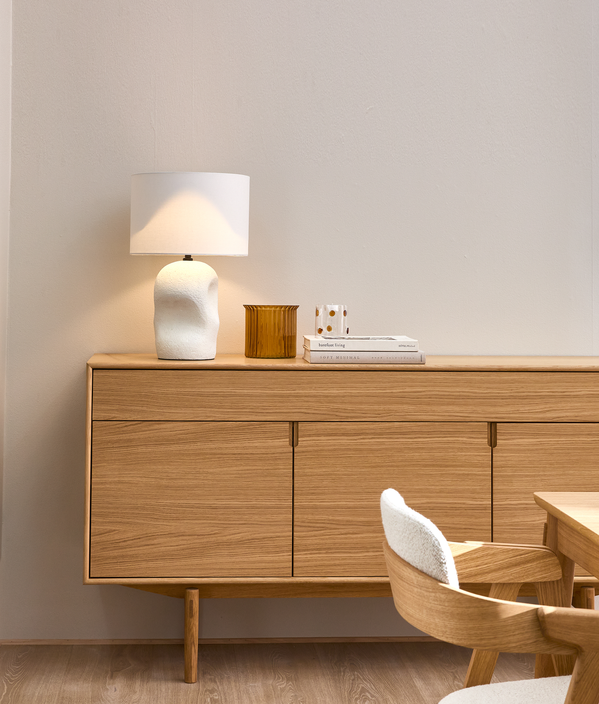 Træskænk med hvid bordlampe, ravfarvet glasbeholder og bøgerne ”hardcover living” og ”soft minimal” i en lys spisestue med god tom vægflade.