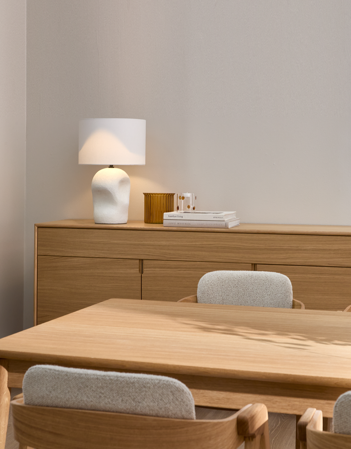 Lyst spisestue med egetræsbord og stole; på skænken står en hvid bordlampe, en ravfarvet vase og en bogstak med “Nordic Living”.