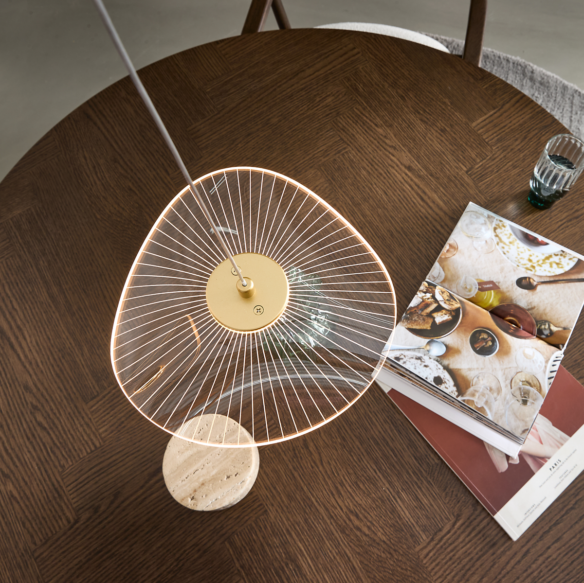 Overhead-billede af rundt spisebord i mørkt træ med moderne bordlampe i glas og messing med marmorfod, et glas vand og åbne magasiner.